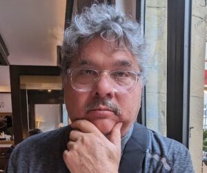 A middle-aged man with gray curly hair, glasses, and a mustache sits indoors, resting his chin on his hand and looking at the camera.
