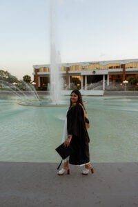A woman in a graduation gown and heels stands by a large fountain with a modern building in the background, holding her cap.