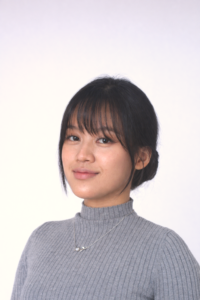 Woman with straight dark hair in a low bun, wearing a gray ribbed turtleneck sweater and a necklace, standing against a plain light background.