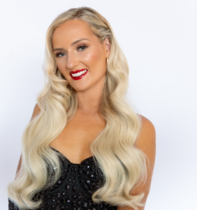 Woman with long wavy blonde hair, red lipstick, and a black sequined top poses in front of a plain white background.