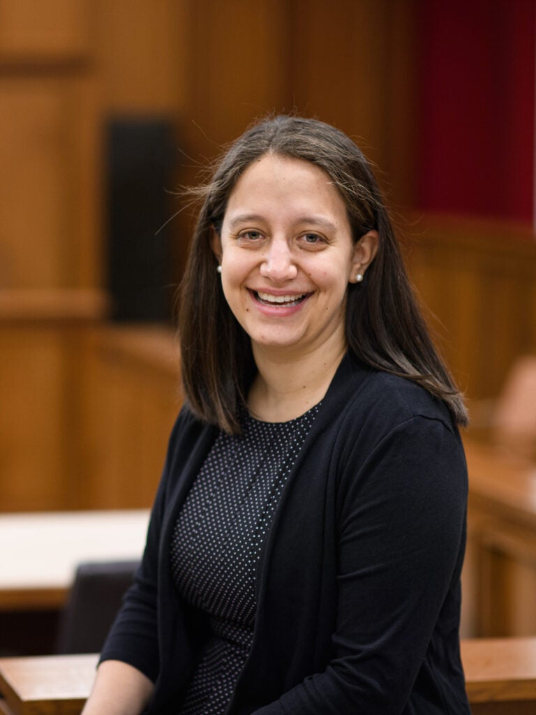 A woman with long dark hair, wearing a black cardigan and polka dot dress, smiles while seated in a wood-paneled courtroom.