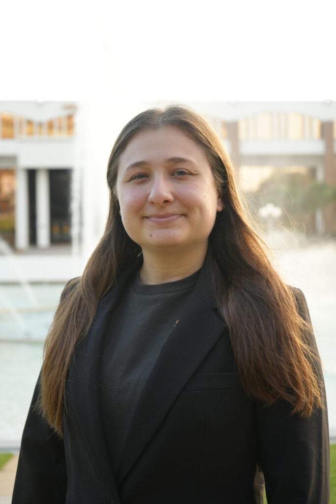 A person with long brown hair, wearing a black blazer and dark shirt, stands outdoors near a fountain and modern buildings.