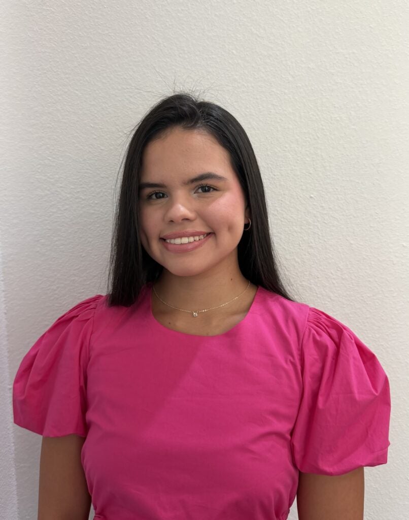 A woman with long dark hair wears a bright pink puff-sleeve top, gold necklace, and smiles in front of a plain white wall.