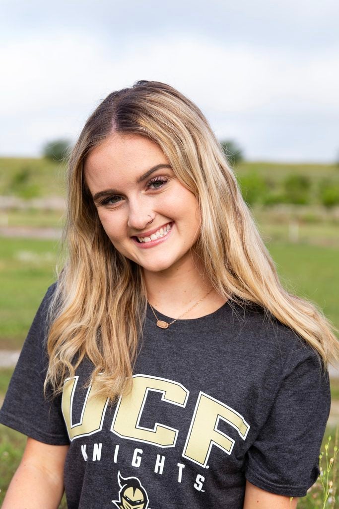 Young woman with long blonde hair smiles outdoors, wearing a dark “UCF Knights” t-shirt. Green grass and a cloudy sky are visible in the background.