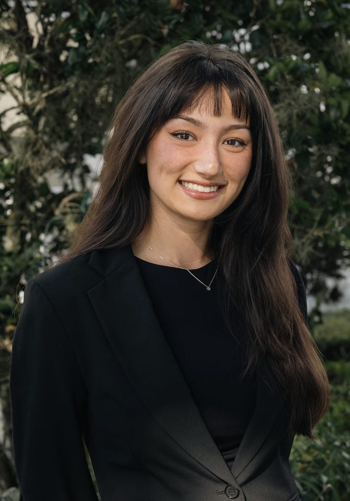 A woman with long brown hair wearing a black blazer and top smiles outdoors, with green foliage in the background.