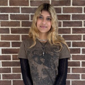 Person with long blonde hair stands in front of a brick wall, wearing a graphic t-shirt and layered necklaces.