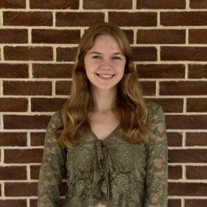 A person with long hair stands against a brick wall, wearing a green lace top and smiling.