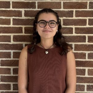 Young woman with glasses and brown sleeveless top stands smiling in front of a brick wall.