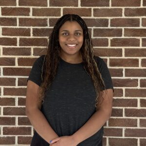 A person with long braided hair smiles, standing in front of a brick wall, wearing a dark t-shirt.