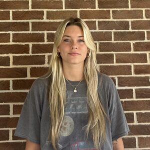 A young woman with long blonde hair wearing a gray graphic T-shirt and light blue jeans stands in front of a brick wall.