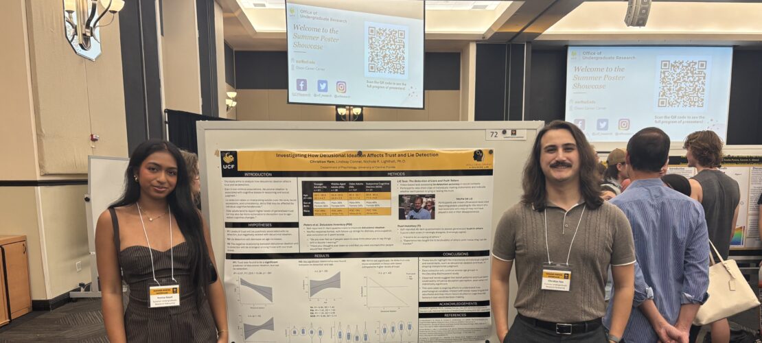 Two people stand beside a scientific poster at a conference. A presentation screen and other attendees are visible in the background.