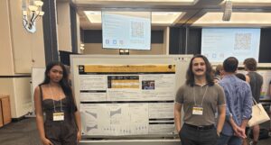 Two people stand beside a scientific poster at a conference. A presentation screen and other attendees are visible in the background.