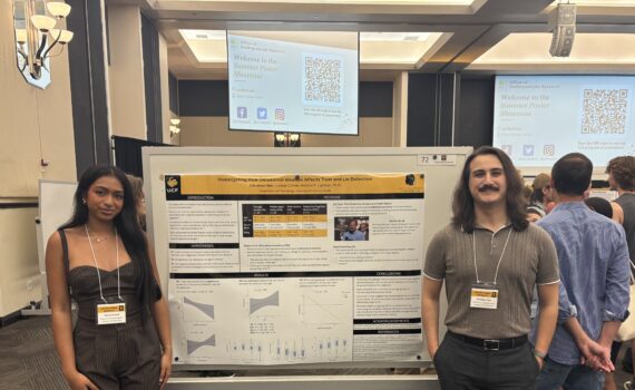 Two people stand beside a scientific poster at a conference. A presentation screen and other attendees are visible in the background.