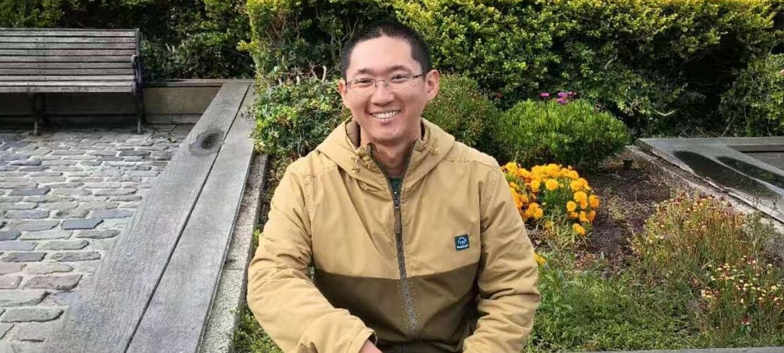 A man wearing glasses and a tan jacket sits on a wooden bench in front of a garden with a large crab sculpture in the background.