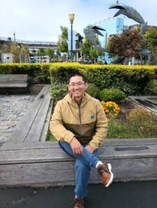 A man wearing glasses and a tan jacket sits on a wooden bench in front of a garden with a large crab sculpture in the background.