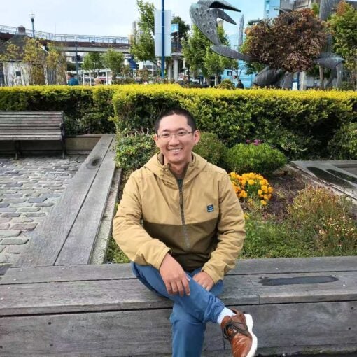 A man wearing glasses and a tan jacket sits on a wooden bench in front of a garden with a large crab sculpture in the background.