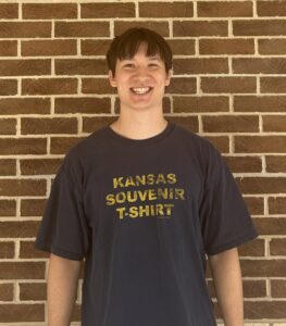 A person stands smiling in front of a brick wall, wearing a dark T-shirt with the text "KANSAS SOUVENIR T-SHIRT" printed in yellow.