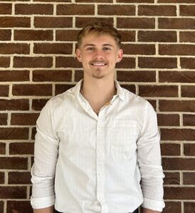 A young man with short blond hair and a goatee, wearing a white button-up shirt, stands smiling in front of a brick wall.