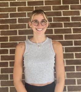 A woman with glasses and a sleeveless gray top stands smiling in front of a brick wall.