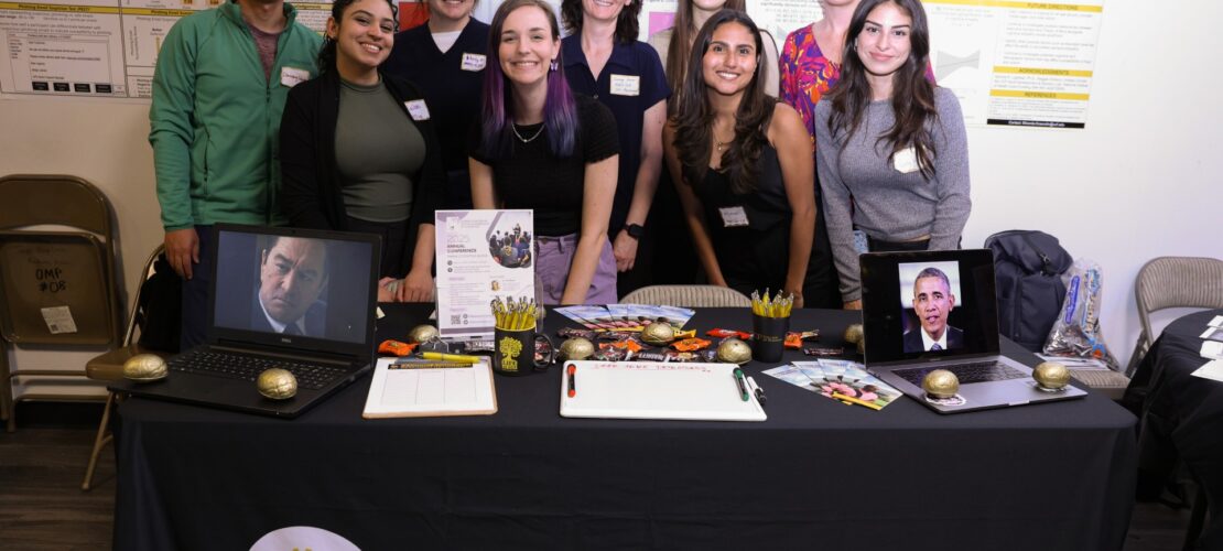 Eight people stand behind a table labeled "Adult Development & Decision Lab" displaying research materials, two framed photos, and various promotional items at a science event.