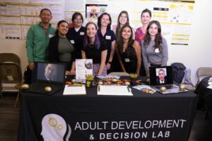 Eight people stand behind a table labeled "Adult Development & Decision Lab" displaying research materials, two framed photos, and various promotional items at a science event.