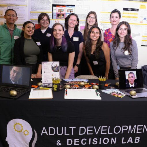 Eight people stand behind a table labeled "Adult Development & Decision Lab" displaying research materials, two framed photos, and various promotional items at a science event.