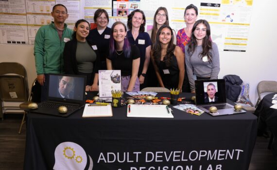 Eight people stand behind a table labeled "Adult Development & Decision Lab" displaying research materials, two framed photos, and various promotional items at a science event.