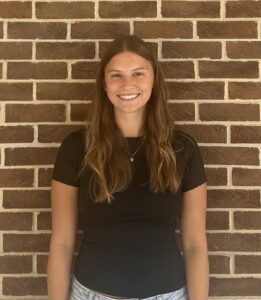 A woman with long brown hair, wearing a black t-shirt and necklace, stands smiling in front of a brick wall.