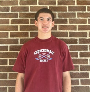 A young person wearing a maroon Abercrombie Hockey t-shirt stands in front of a brick wall, facing the camera and smiling.