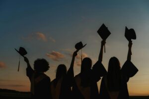 Four graduates in caps and gowns hold their mortarboards in the air at sunset, celebrating their achievement.