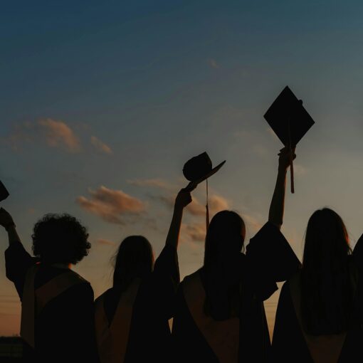 Four graduates in caps and gowns hold their mortarboards in the air at sunset, celebrating their achievement.