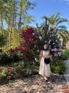 A woman in a cream dress and graduation cap stands in a garden with colorful plants and flowers under a clear blue sky.