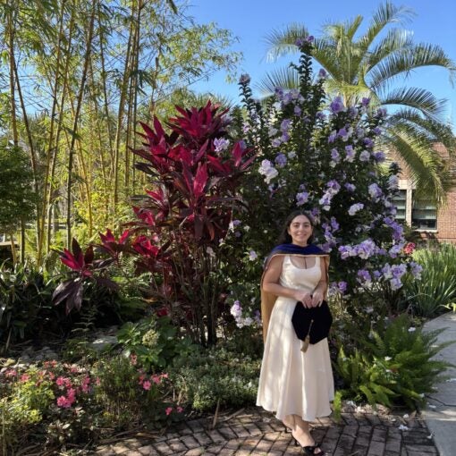A woman in a cream dress and graduation cap stands in a garden with colorful plants and flowers under a clear blue sky.
