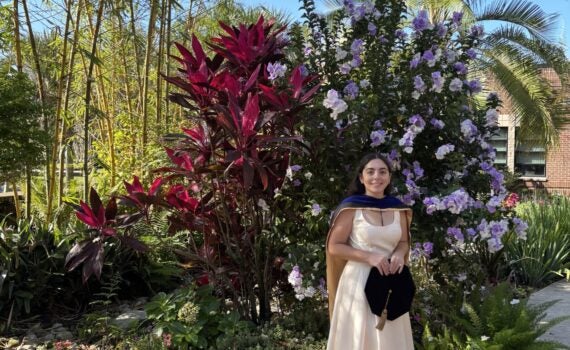 A woman in a cream dress and graduation cap stands in a garden with colorful plants and flowers under a clear blue sky.