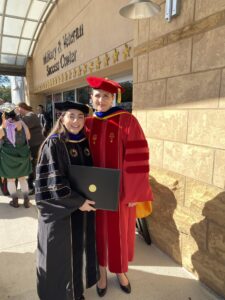 Two women in academic regalia pose outside the Military & Veteran Success Center; one holds a diploma and both are smiling at the camera.