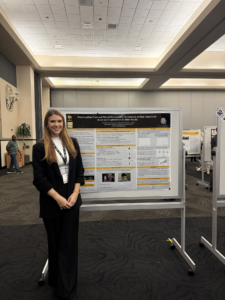 A woman stands smiling in front of a scientific poster display in a large conference room with high ceilings and neutral tones.