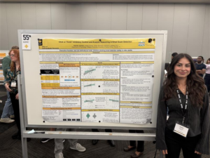 A woman stands next to a scientific research poster titled "Click or Think? Inhibitory Control and Analytic Reasoning in Email Scam Detection" at a conference, with attendees in the background.