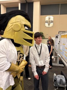 A person stands next to a UCF mascot in front of a research poster at an indoor event with other attendees in the background.