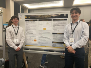 Two men in dress shirts stand on either side of a research poster at a conference, with a UCF logo visible on the poster behind them.