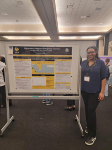 A woman stands next to a scientific research poster titled "Risk Sensitivity and Decision Making Under Uncertainty" at an indoor academic conference.