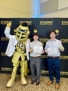 Two people hold certificates and awards while posing with the UCF knight mascot in front of a "Student Research Week" backdrop.