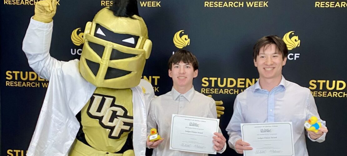 Two people hold certificates and awards while posing with the UCF knight mascot in front of a "Student Research Week" backdrop.