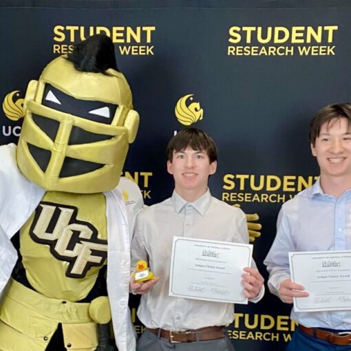 Two people hold certificates and awards while posing with the UCF knight mascot in front of a "Student Research Week" backdrop.