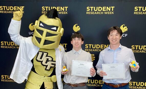 Two people hold certificates and awards while posing with the UCF knight mascot in front of a "Student Research Week" backdrop.