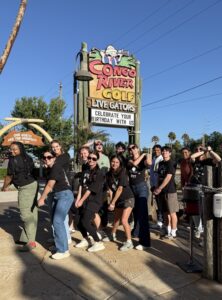 A group of people poses in front of the Cow Creek's River Adventure sign at an outdoor attraction on a sunny day.