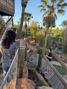 People play mini-golf on a course with wooden bridges, palm trees, and rocks under a clear blue sky.