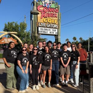 A group of people wearing matching black shirts stand together in front of a Congo River Golf sign on a sunny day.