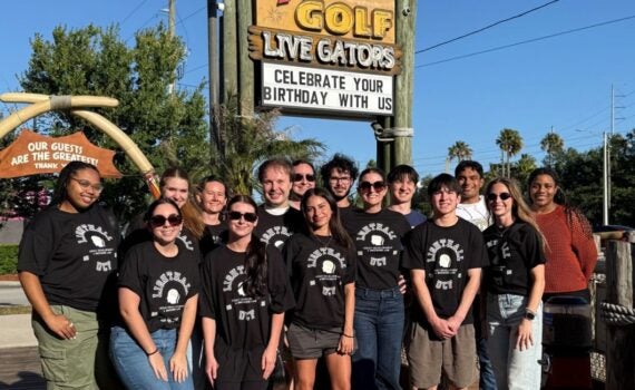 A group of people wearing matching black shirts stand together in front of a Congo River Golf sign on a sunny day.