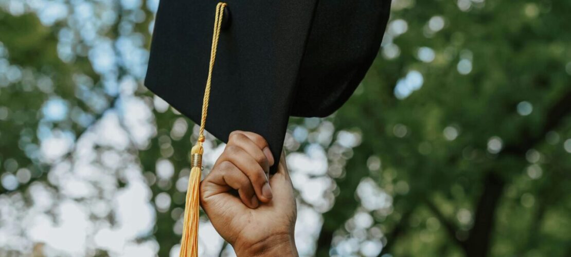 A person in a graduation gown holds up a graduation cap with a yellow tassel outdoors, with trees in the background.