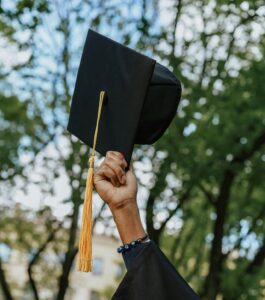 A person in a graduation gown holds up a graduation cap with a yellow tassel outdoors, with trees in the background.